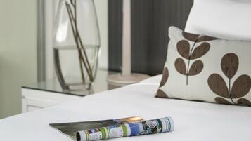 Una cama bien hecha con un cojín, una revista, un florero con flores y una lámpara.