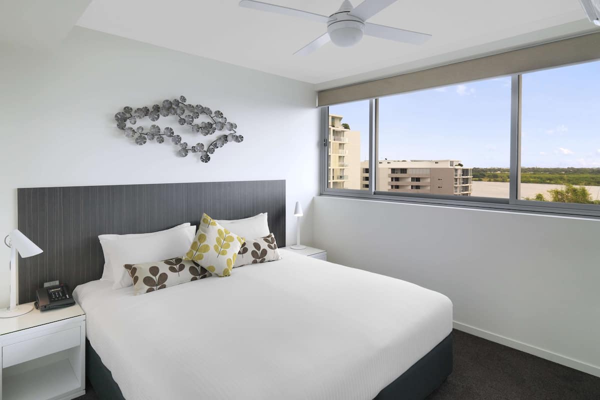 A hotel room with a large bed, a ceiling fan, a window with a view of buildings, and a decorative wall piece.