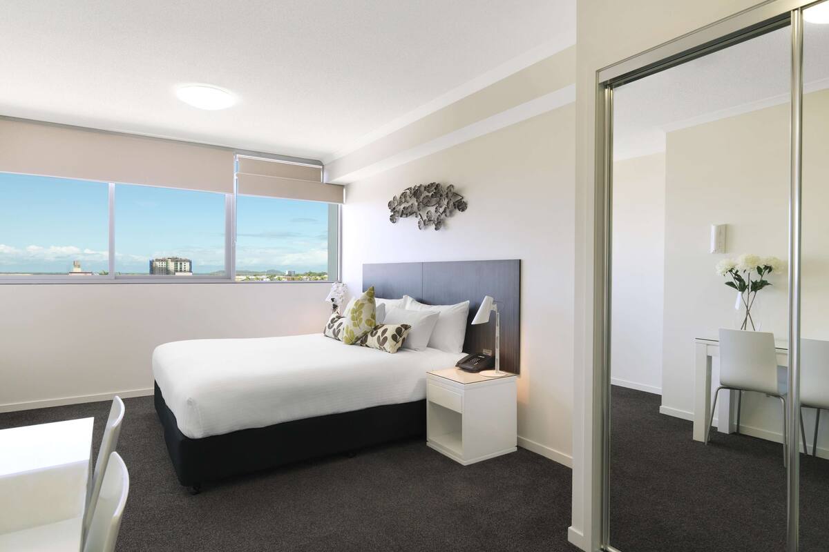 A modern hotel room with a large bed, bedside table, lamp, and a view of the cityscape through the window.