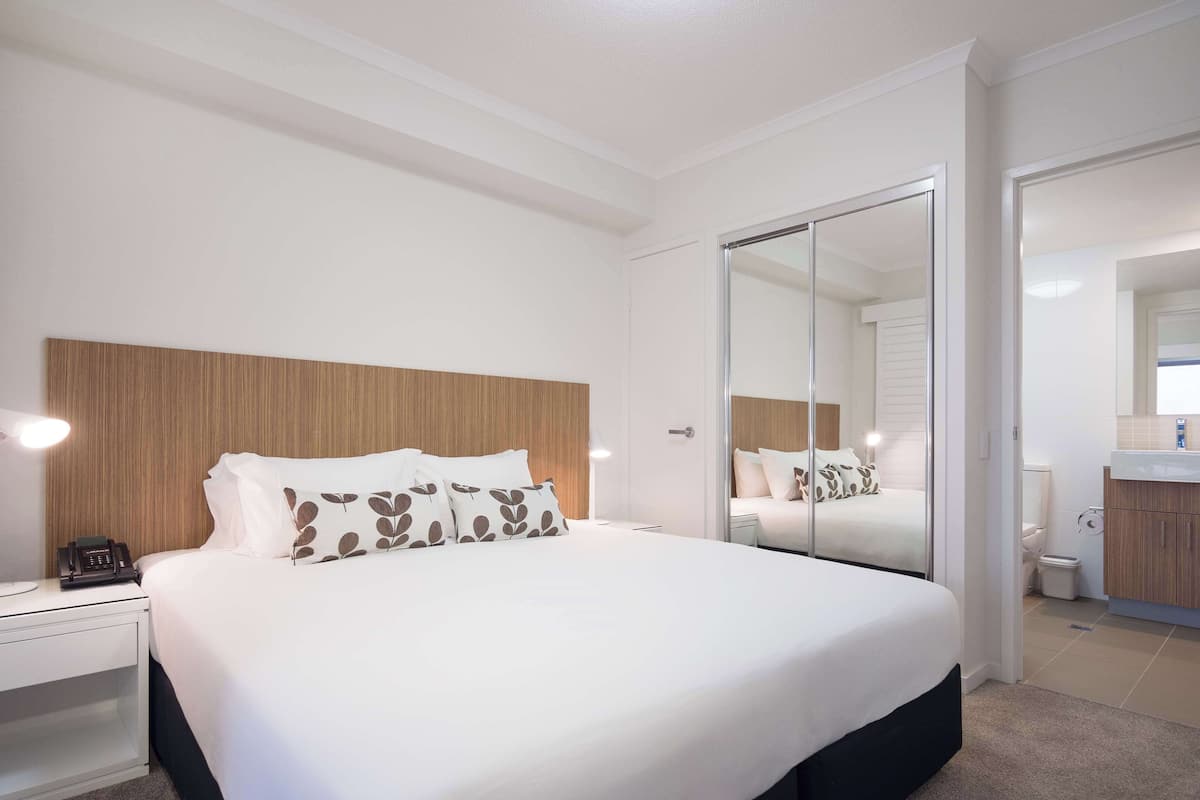 A modern hotel room with a large bed, a wooden headboard, a bedside table with a lamp, and a bathroom visible through a sliding door.
