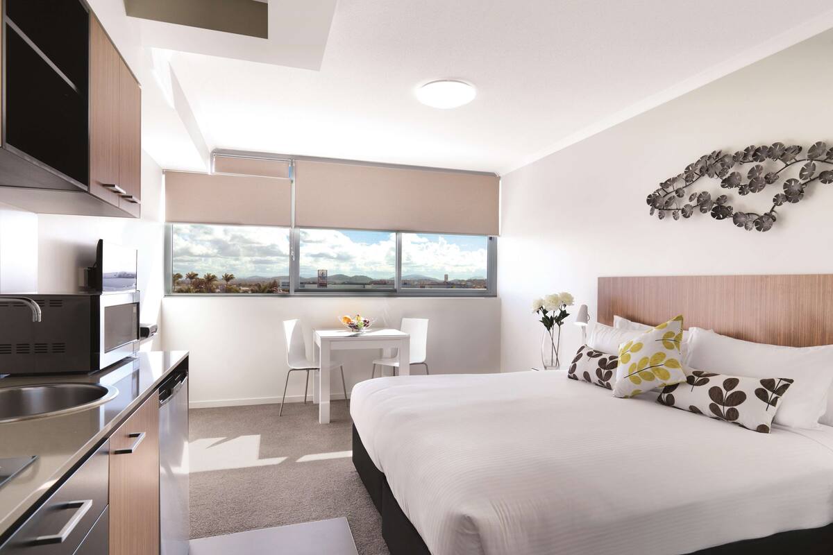A modern hotel room with a bed, a small table with chairs, a kitchenette, and a large window offering a city view.