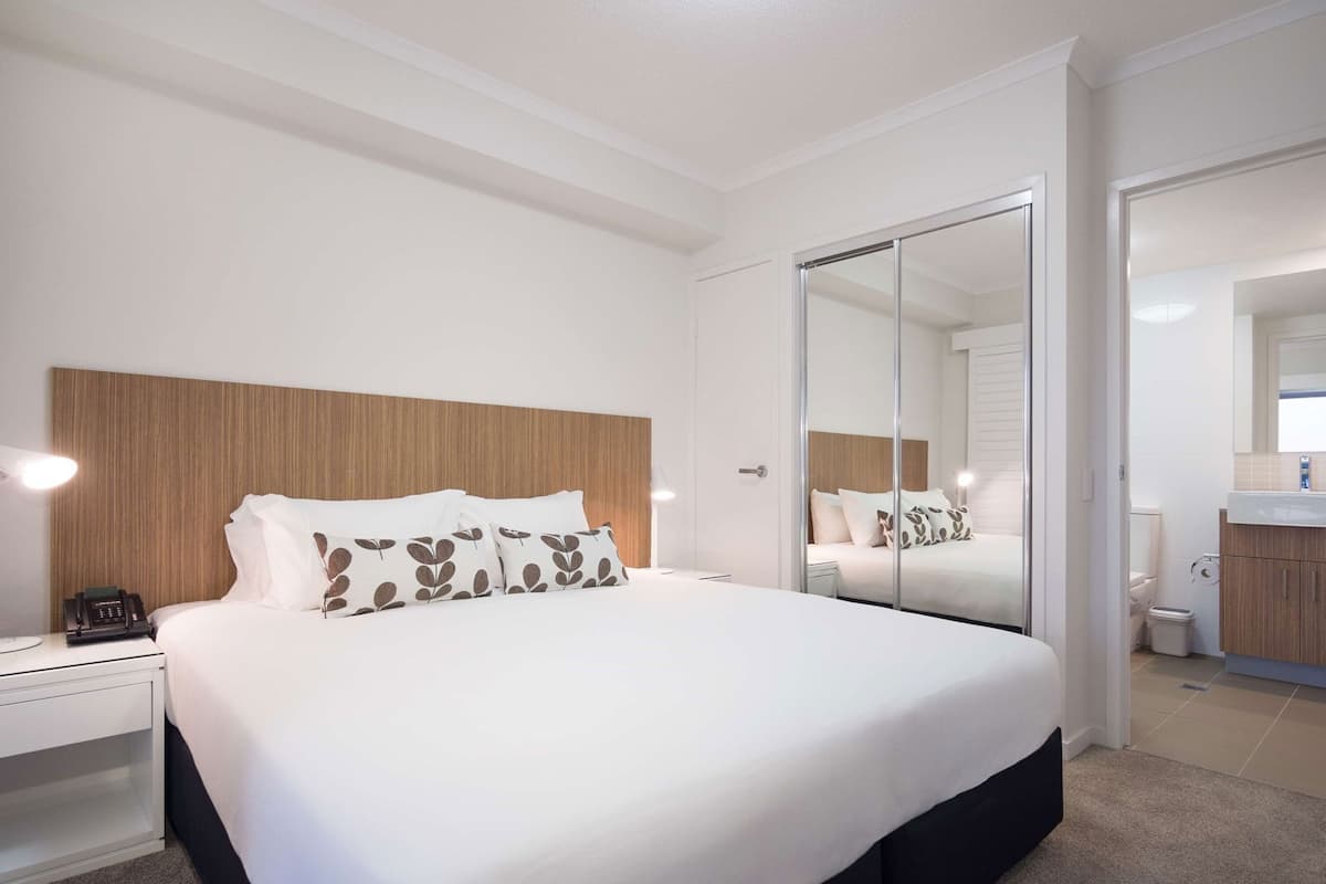 A modern hotel room with a large bed, a bedside telephone, a mirror, and a bathroom visible through an open door.