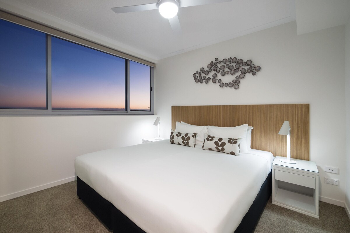 A modern bedroom with a large bed, a ceiling fan, a nightstand, and a wall-mounted decorative piece.