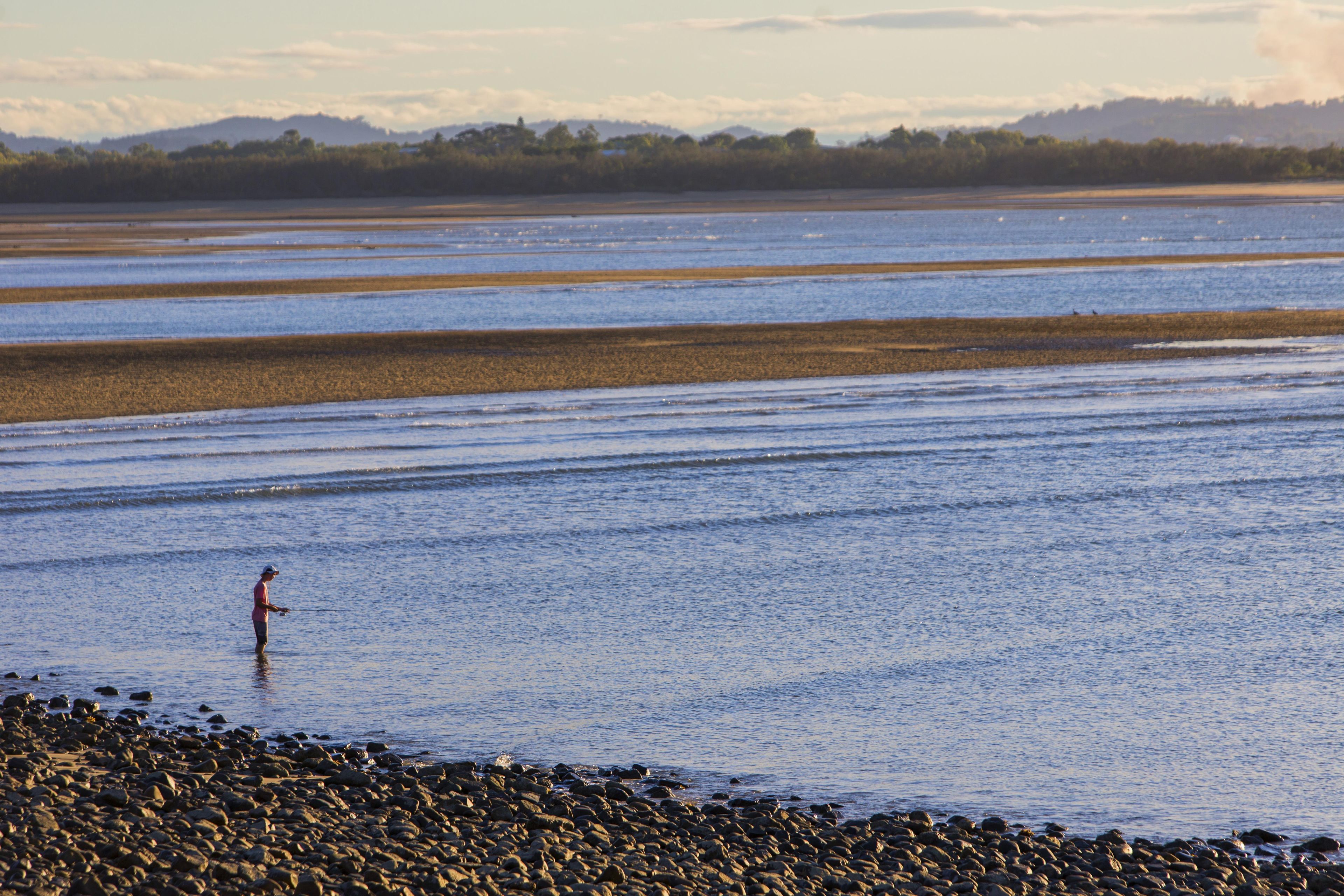 A wide river with a person fishing, surrounded by a rocky shoreline and distant hills.
