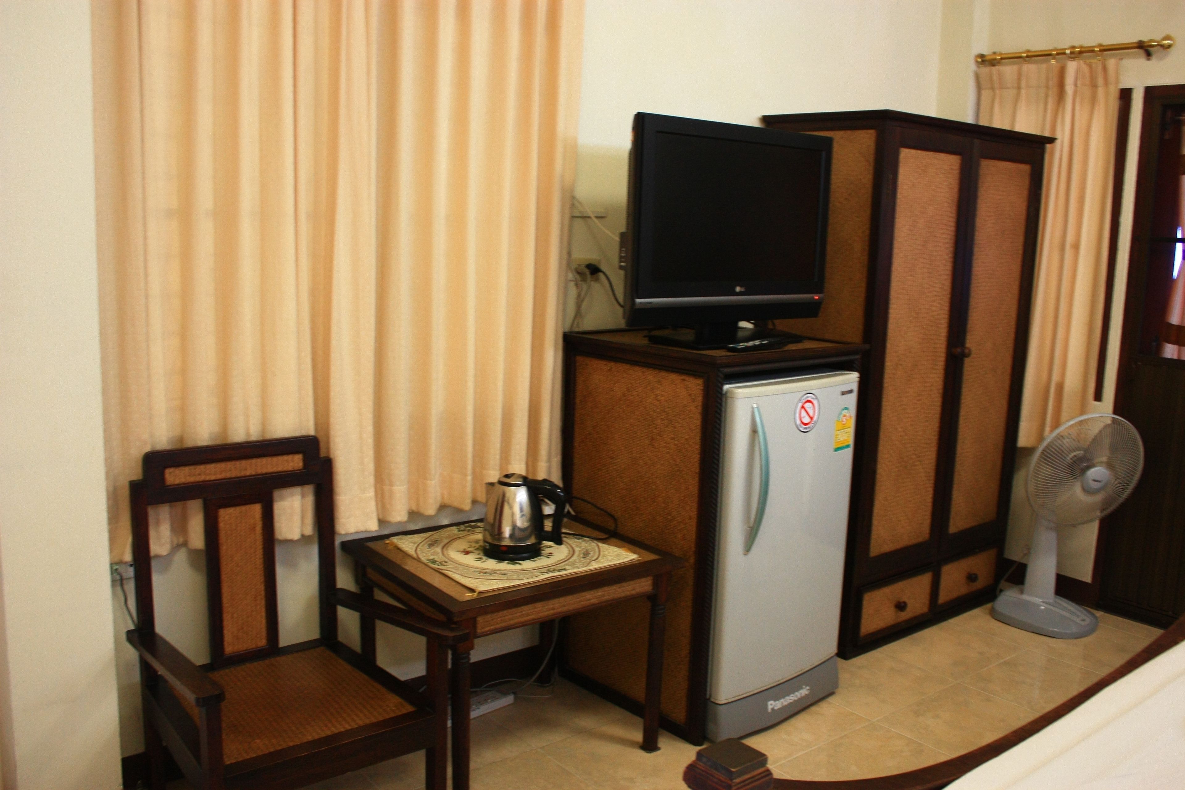 A compact hotel room with a wooden chair, a small table with a kettle, a refrigerator, a television on top of the refrigerator, and a standing fan.