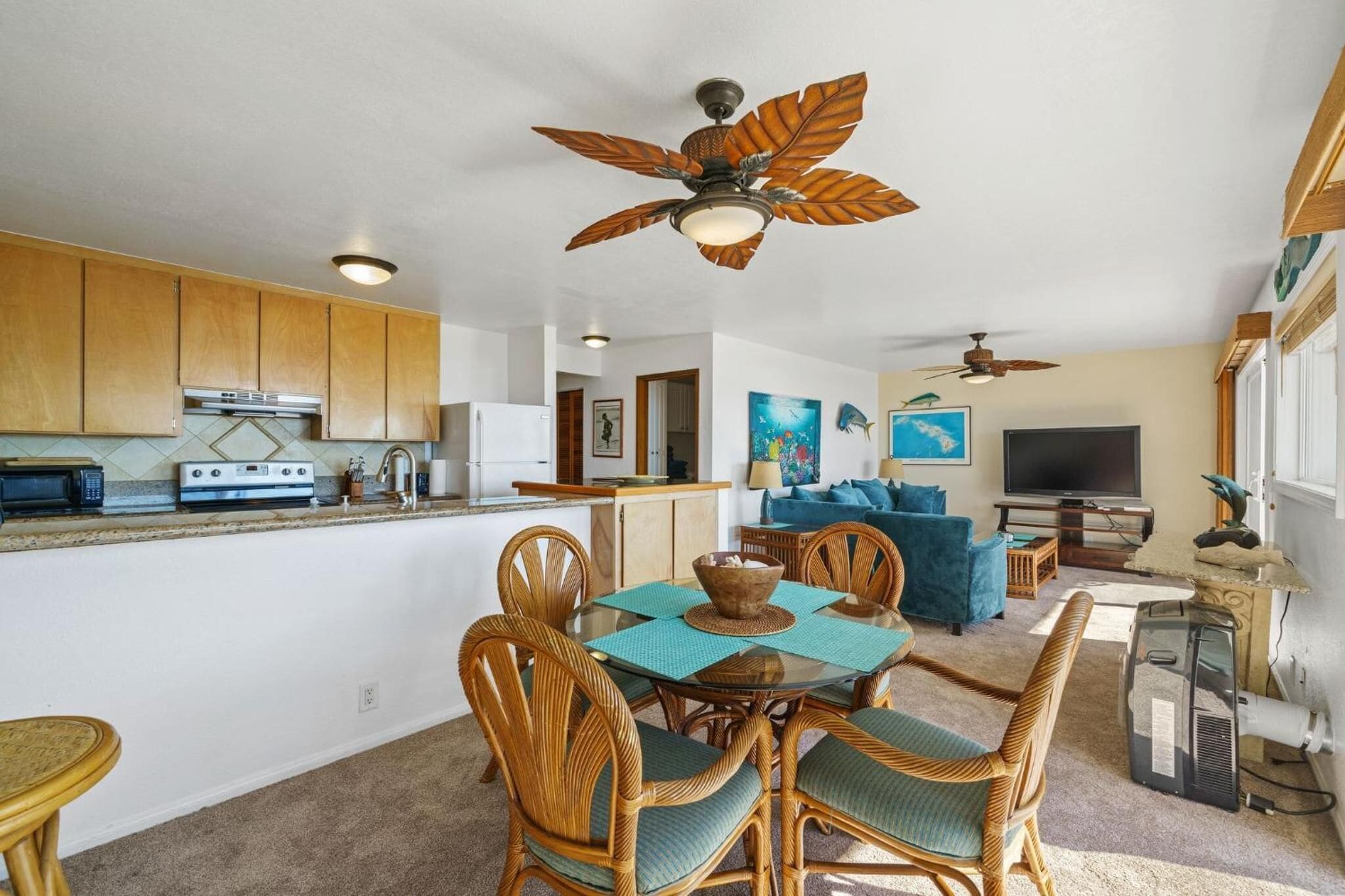 A spacious living area with a dining table, wicker chairs, a ceiling fan, a television, and a kitchen with wooden cabinets and appliances.