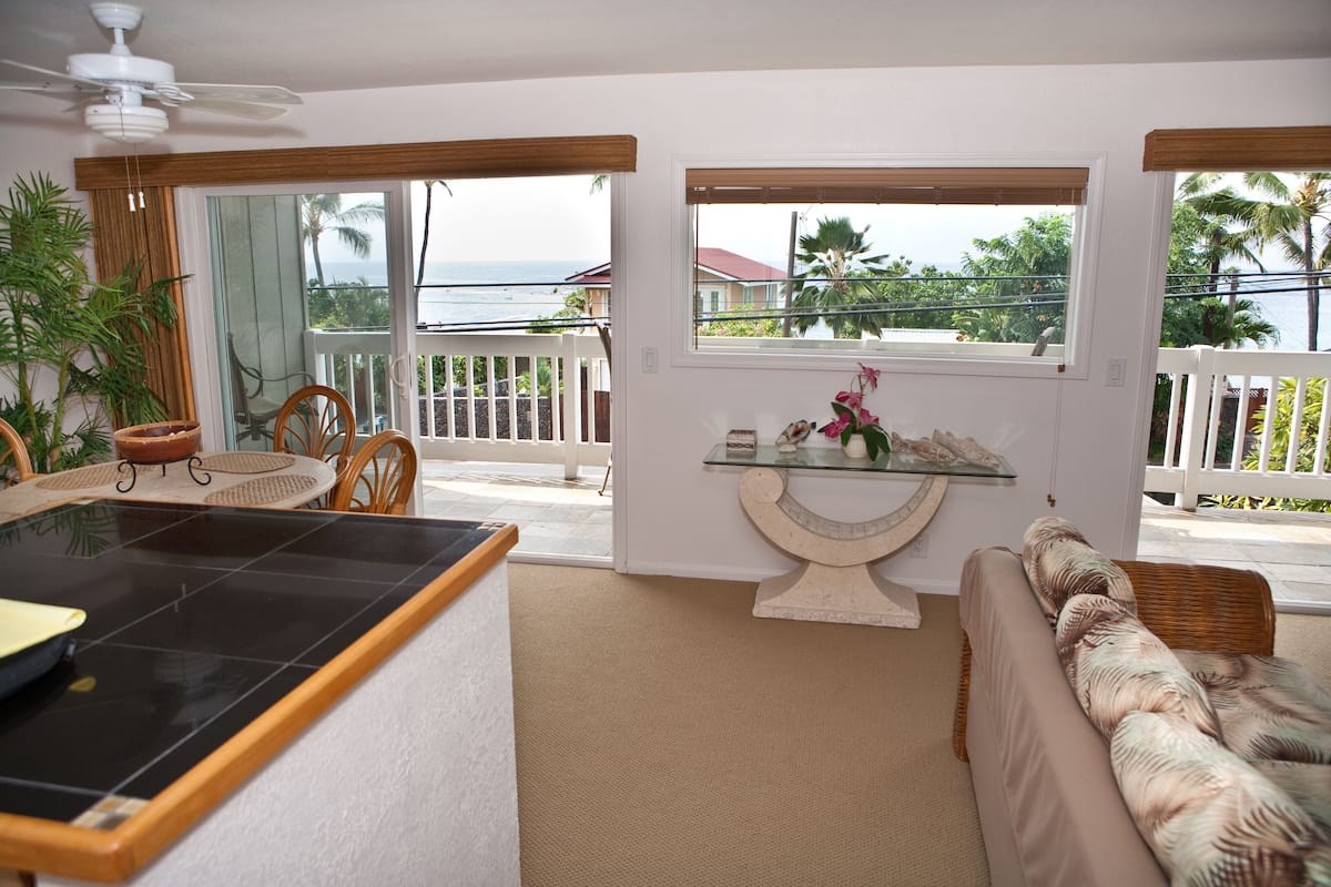 A spacious living area with a kitchen counter, a sofa with patterned cushions, a dining table with chairs, and a view of the ocean through large windows.