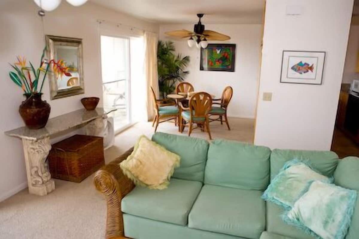 A living room with a green sofa, a dining area with wooden chairs, a ceiling fan, and a painting on the wall.