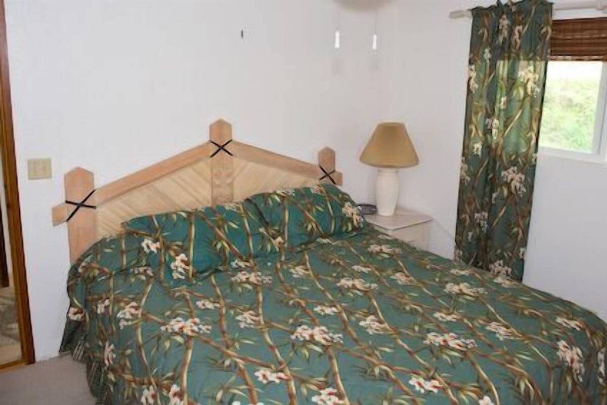 A bedroom with a wooden headboard, floral bedding, a bedside lamp, and a window with curtains.