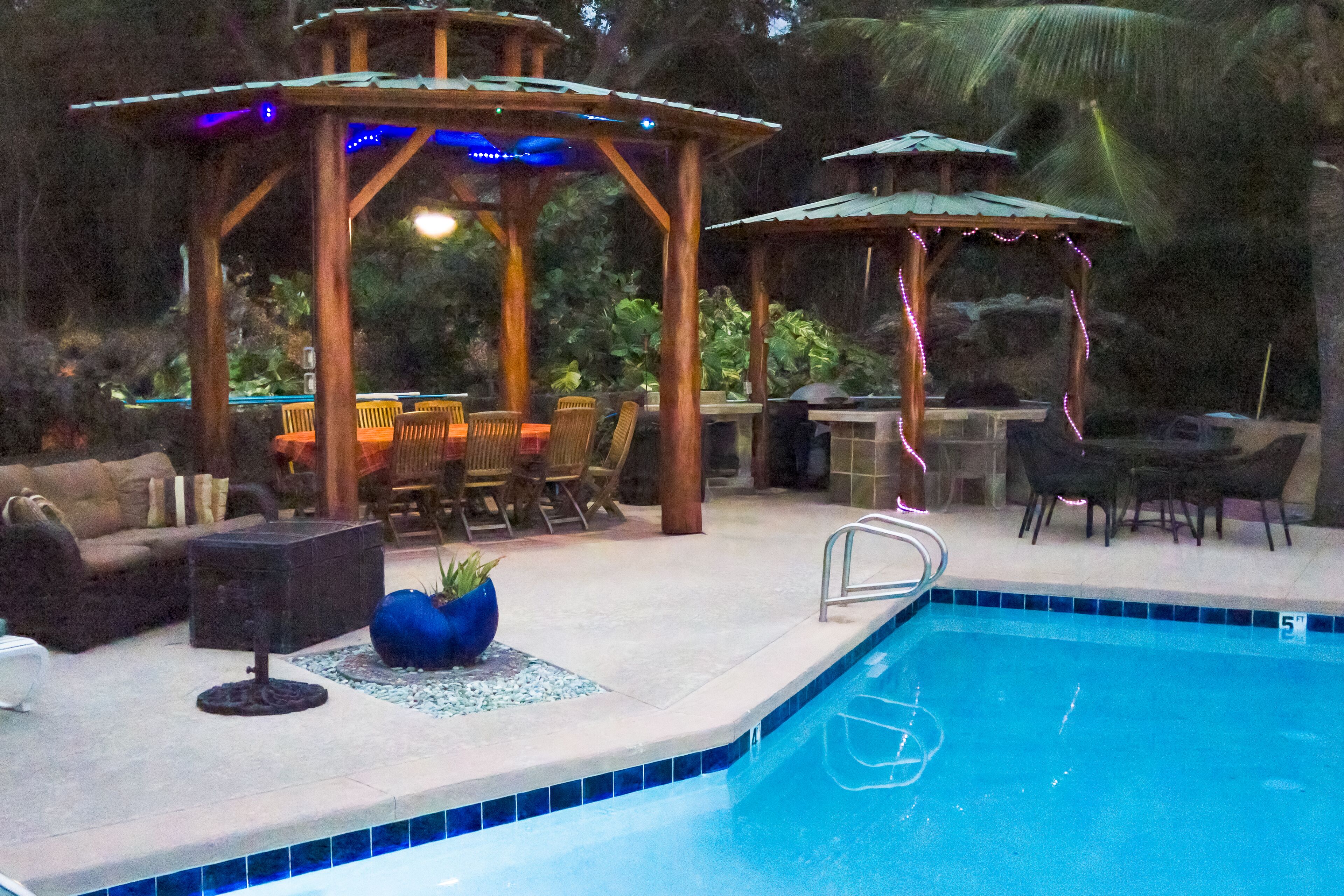 An outdoor pool area with a wooden gazebo, lounge chairs, and a dining table.