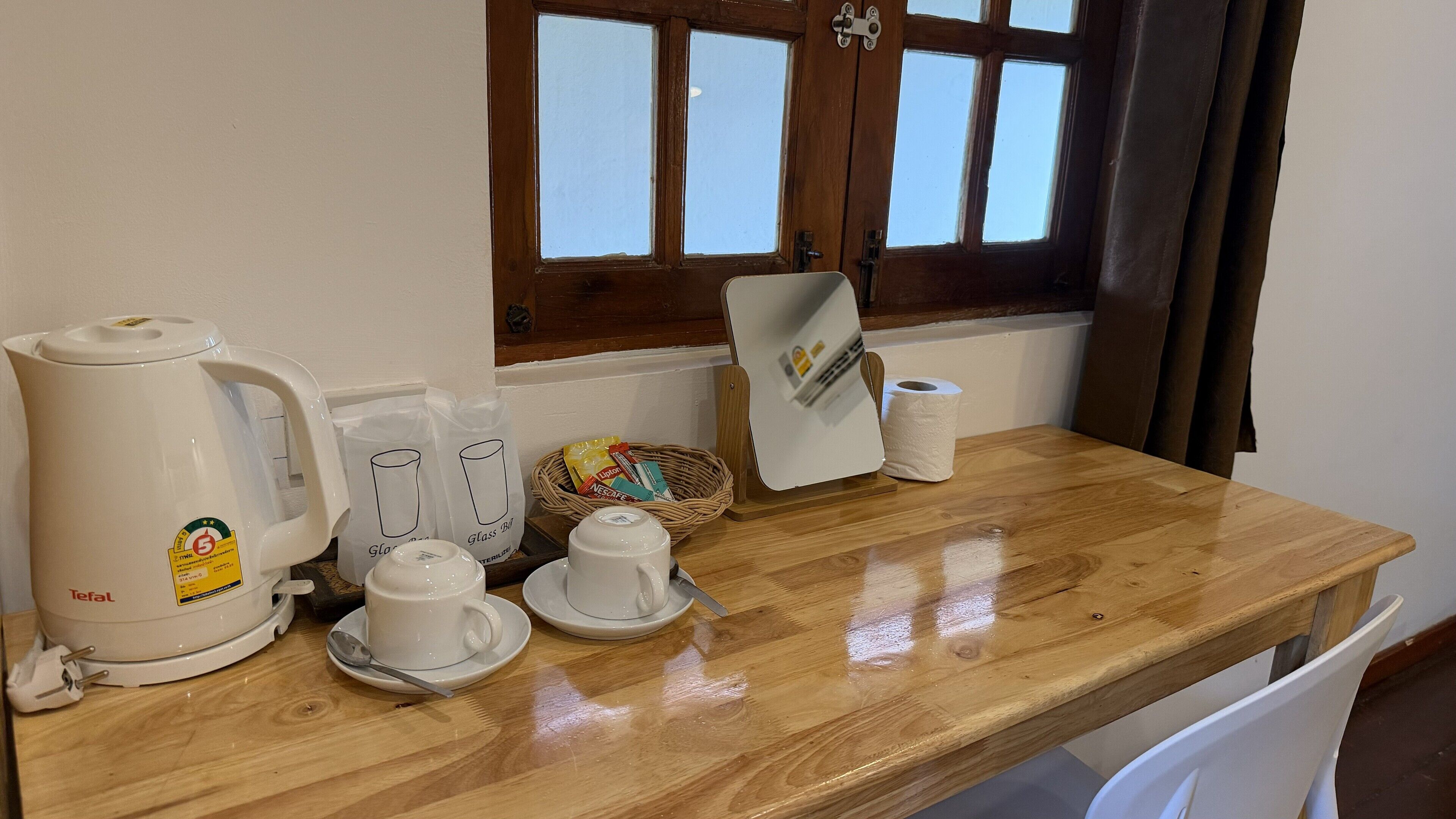 A wooden table with a coffee maker, cups, a basket with snacks, and a window with curtains.
