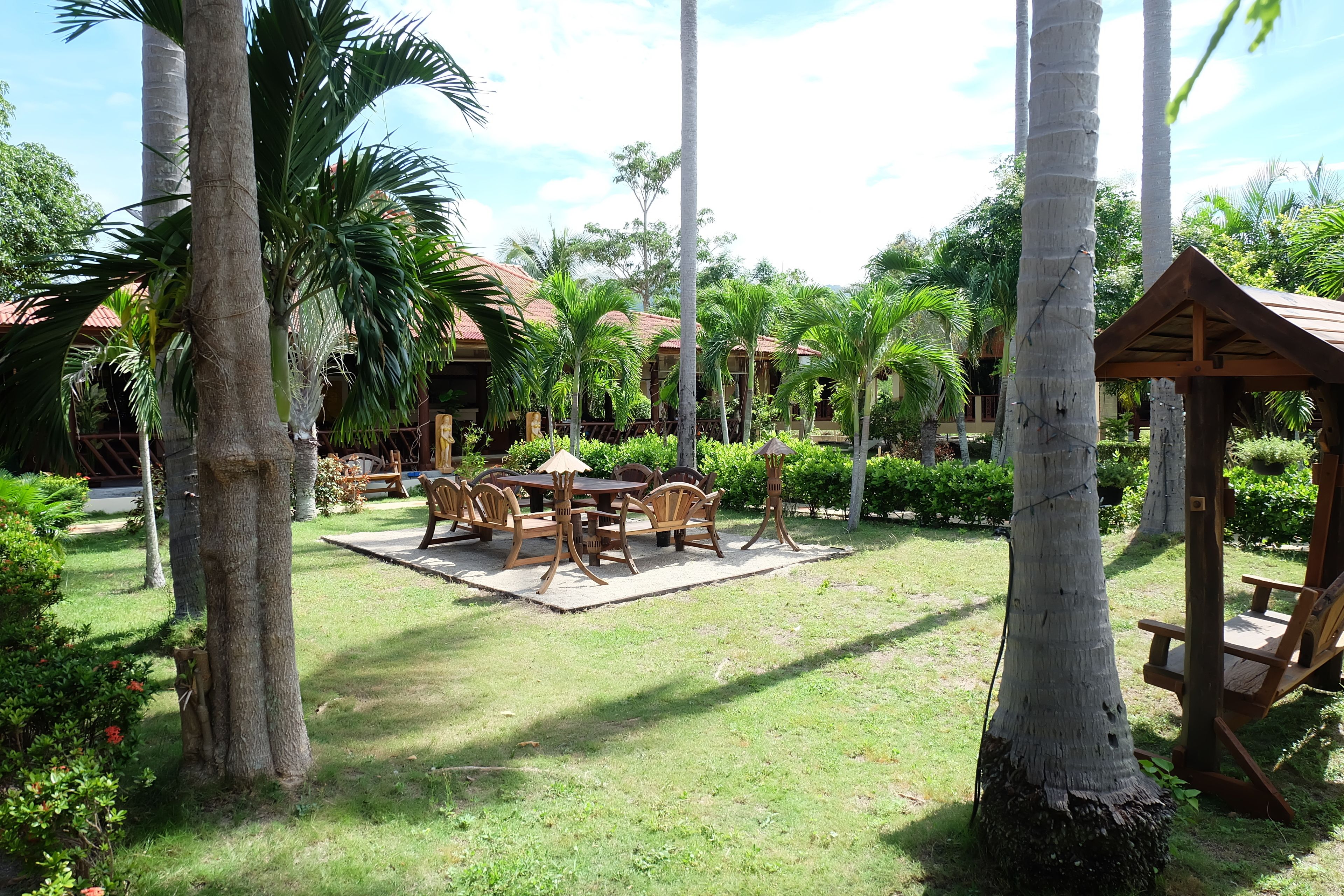 A garden area with a wooden table and chairs, surrounded by palm trees and a gazebo.