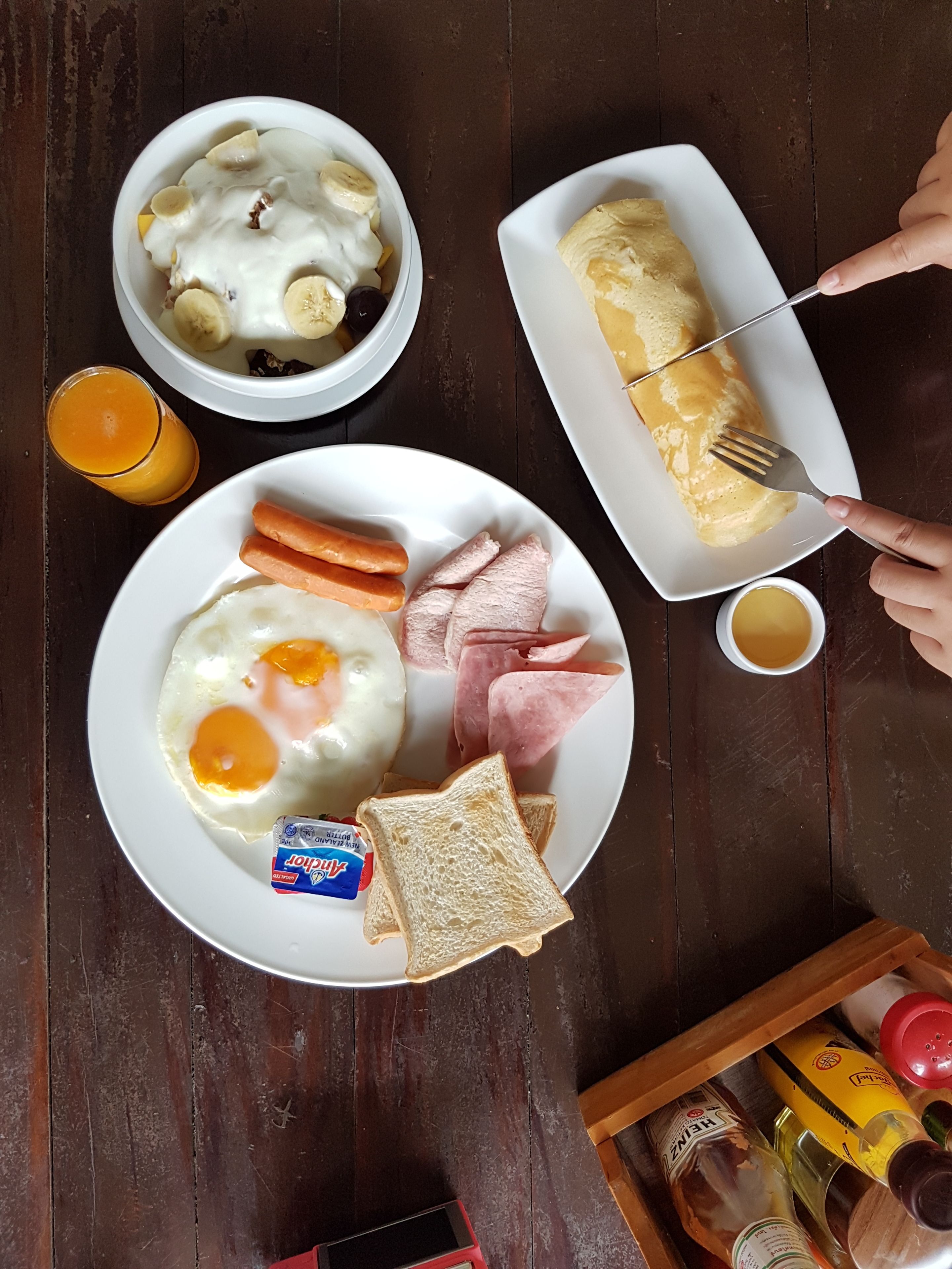 A breakfast plate with eggs, ham, and toast, a bowl of cereal with bananas, and a glass of orange juice on a wooden table.