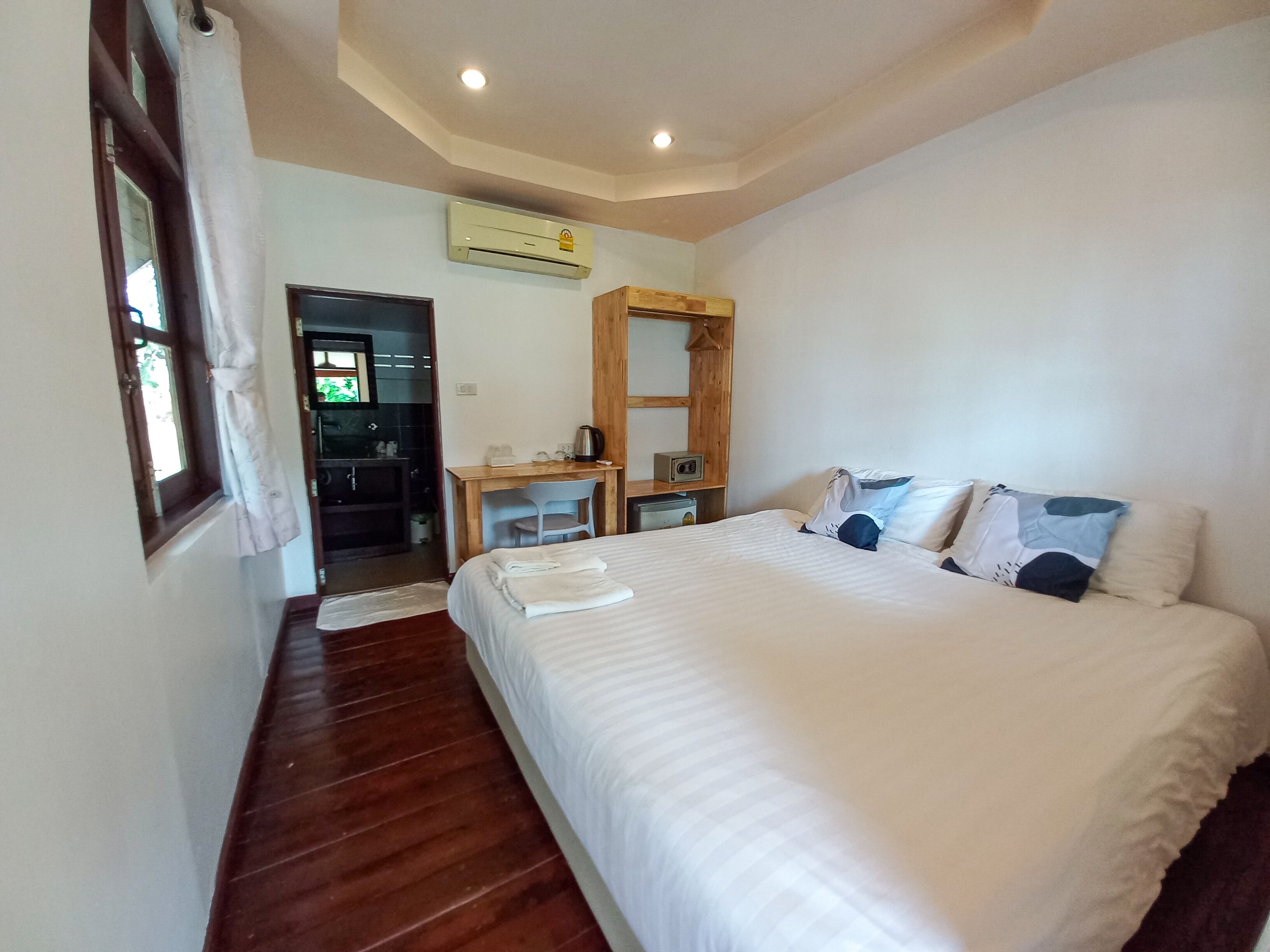 A bedroom with a large bed, a wooden desk, a chair, a small table with a kettle, and an air conditioning unit on the wall.