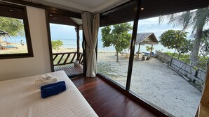 Een kamer met een groot raam met uitzicht op een strand, een bed met handdoeken en een houten terras.