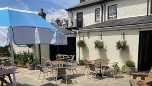 An outdoor patio area with wooden tables and chairs, a blue umbrella, and hanging flower baskets.