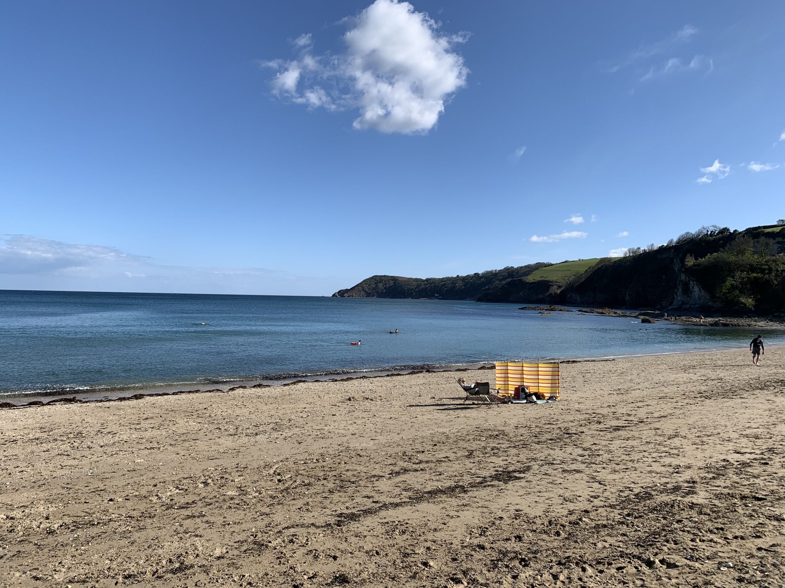 A beach with a sandy shore, a striped beach umbrella, and a person walking.