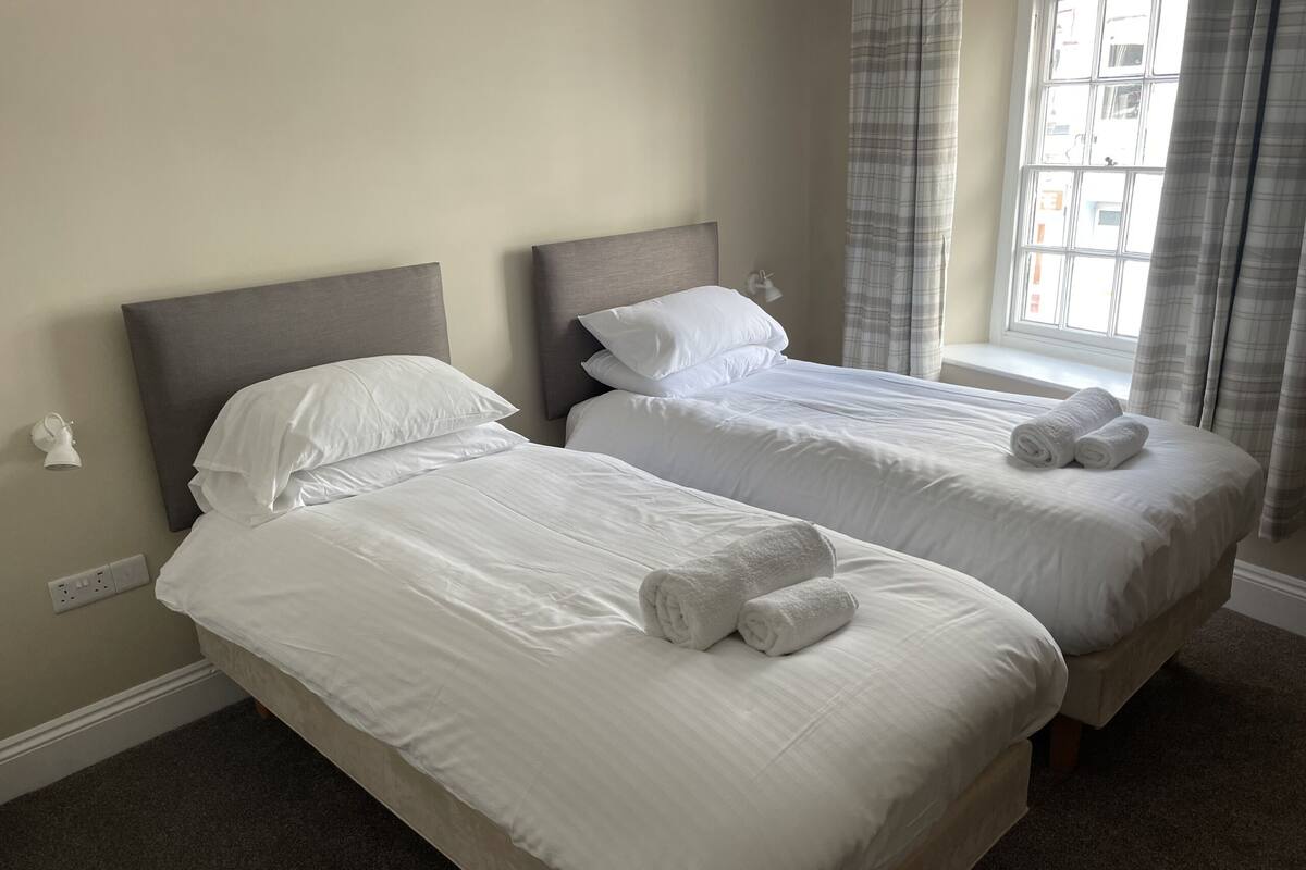 Une chambre d’hôtel avec deux lits simples, du linge de lit blanc, une lampe de chevet et une fenêtre avec des rideaux.