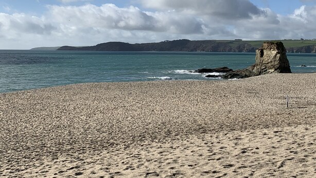 A pebbly beach with a prominent rock formation and distant hills.