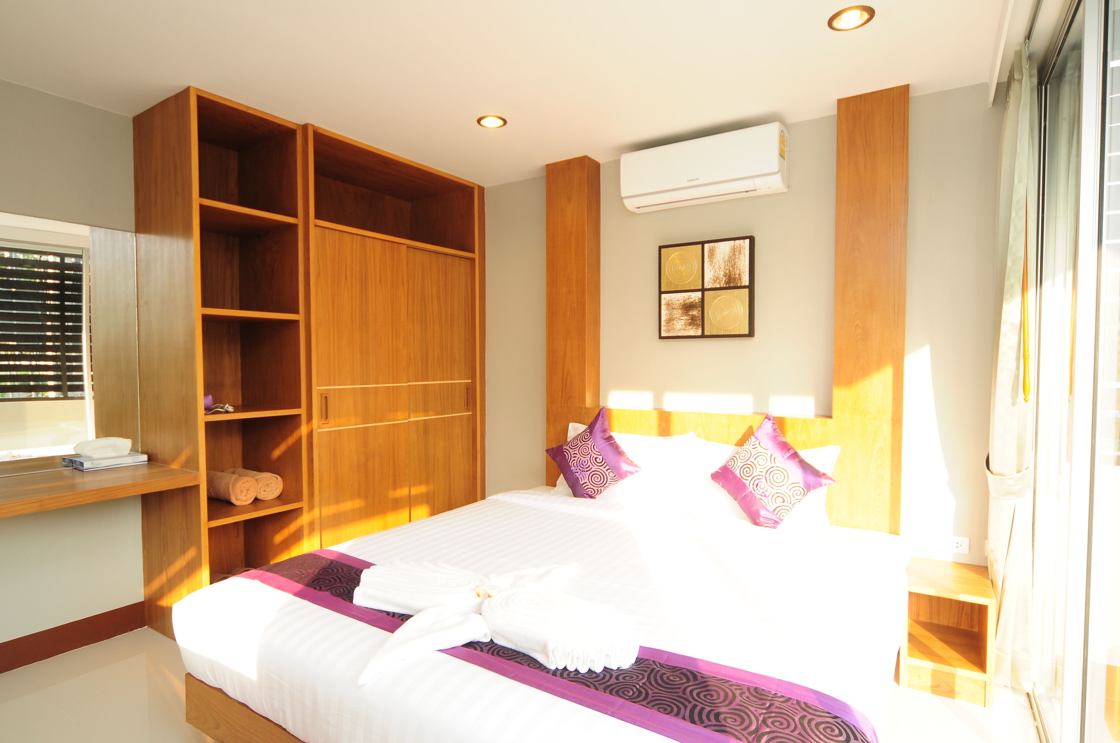 A hotel room with a large bed, wooden wardrobe, built-in shelves, a bedside table, and a window with blinds.