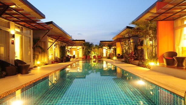 A swimming pool with tiled flooring, surrounded by lounge chairs and potted plants, with buildings on either side.