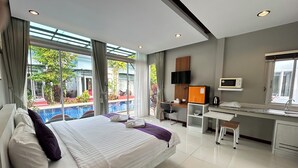 A modern hotel room with a large bed, a desk, a chair, a TV, a microwave, and a view of a swimming pool and greenery.