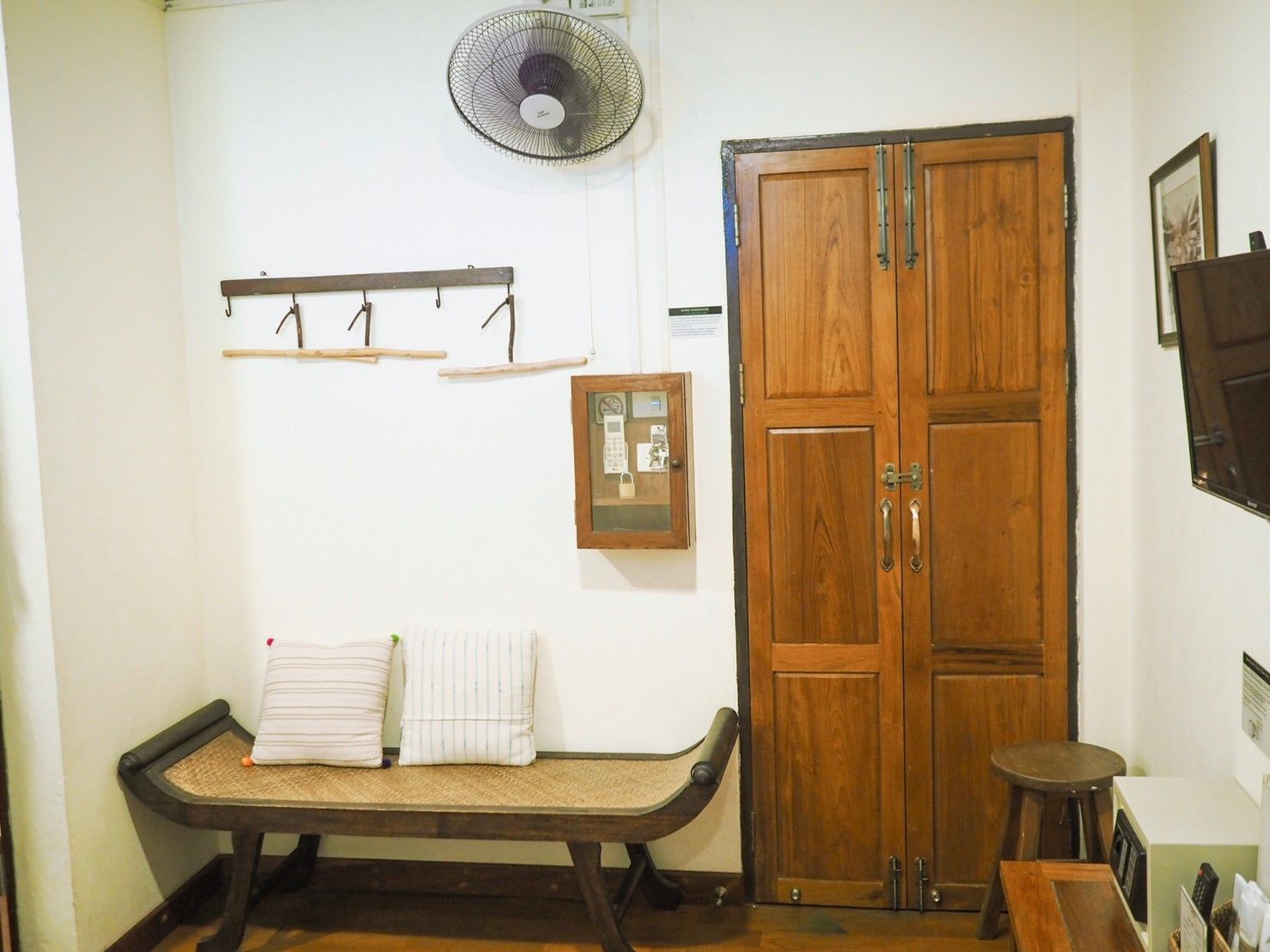 A room with a wooden bench, a wooden wardrobe, a wall-mounted fan, and a small stool.