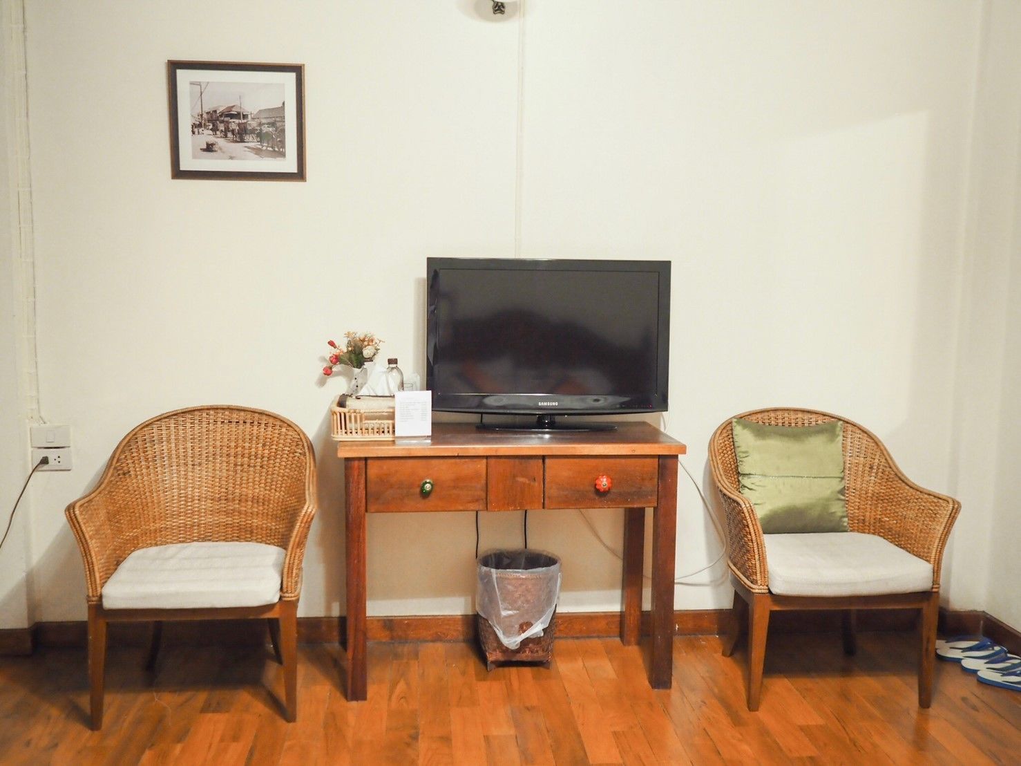 A room with a wooden desk, two wicker chairs, a television, and a trash bin.