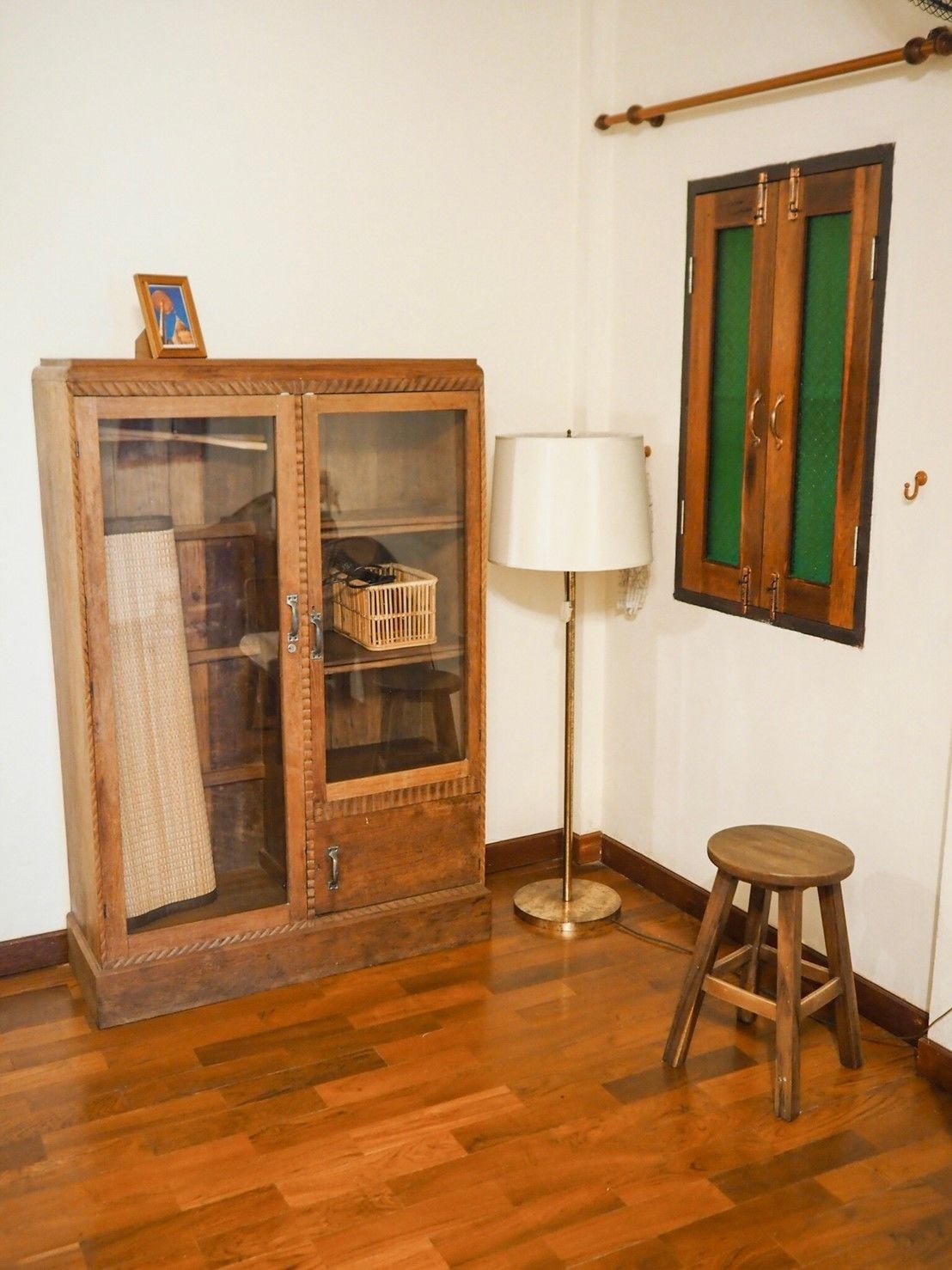 A wooden cabinet with glass doors, a floor lamp, and a stool in a room with wooden flooring.