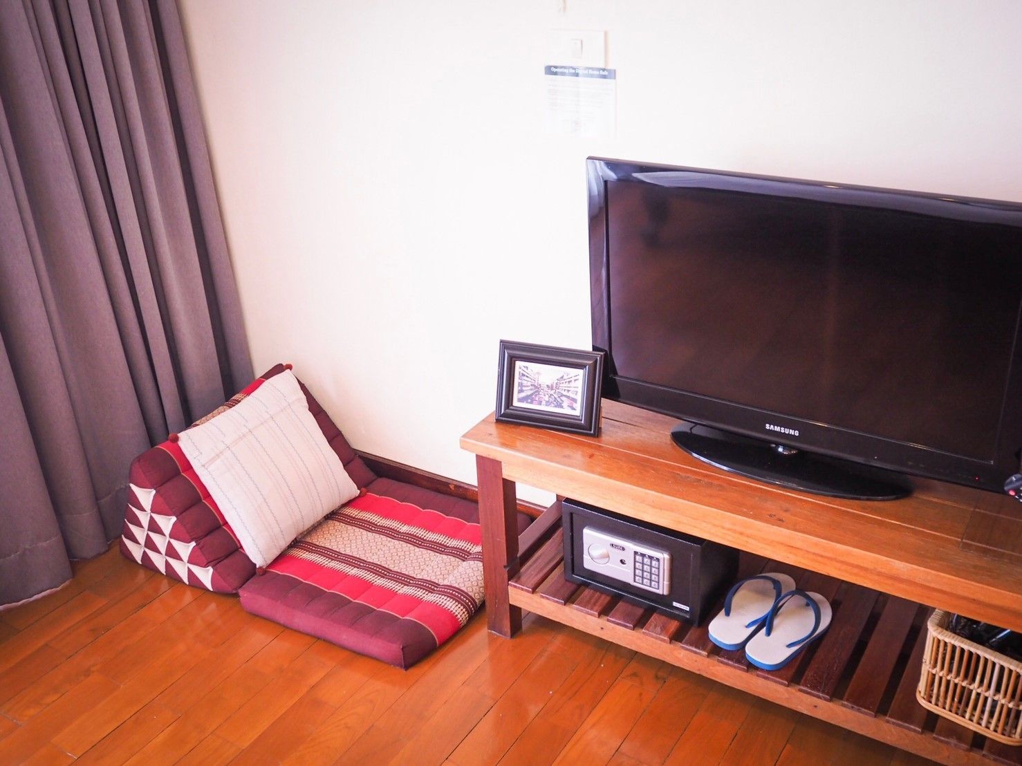 A living room with a flat-screen TV on a wooden stand, a cushioned sofa with a patterned throw, a small framed picture, a remote control, and a pair of flip-flops on the floor.