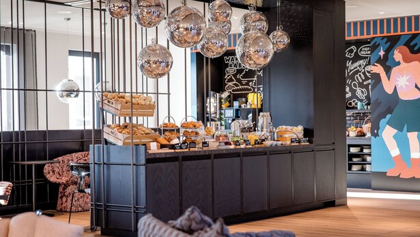 A modern cafe with a central counter displaying pastries and beverages, surrounded by seating areas and decorative disco balls. - Motel One Berlin-Potsdamer Platz (Berlin)
