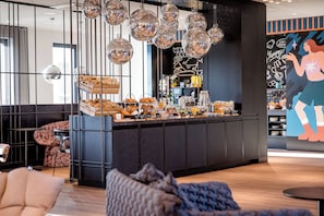 A modern cafe with a central counter displaying pastries and beverages, surrounded by seating areas and decorative disco balls. - Motel One Berlin-Potsdamer Platz (Berlin)