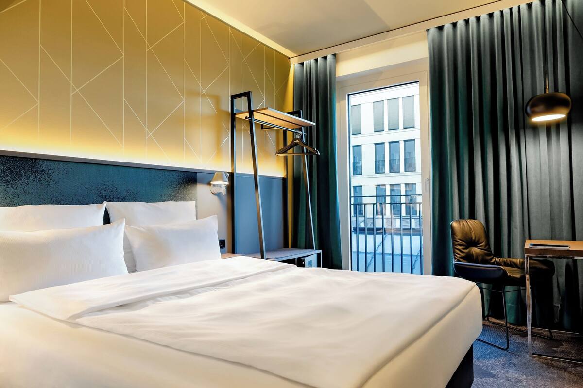 A hotel room with a large bed, a desk, and a chair. There is a window with curtains and a view of buildings outside.