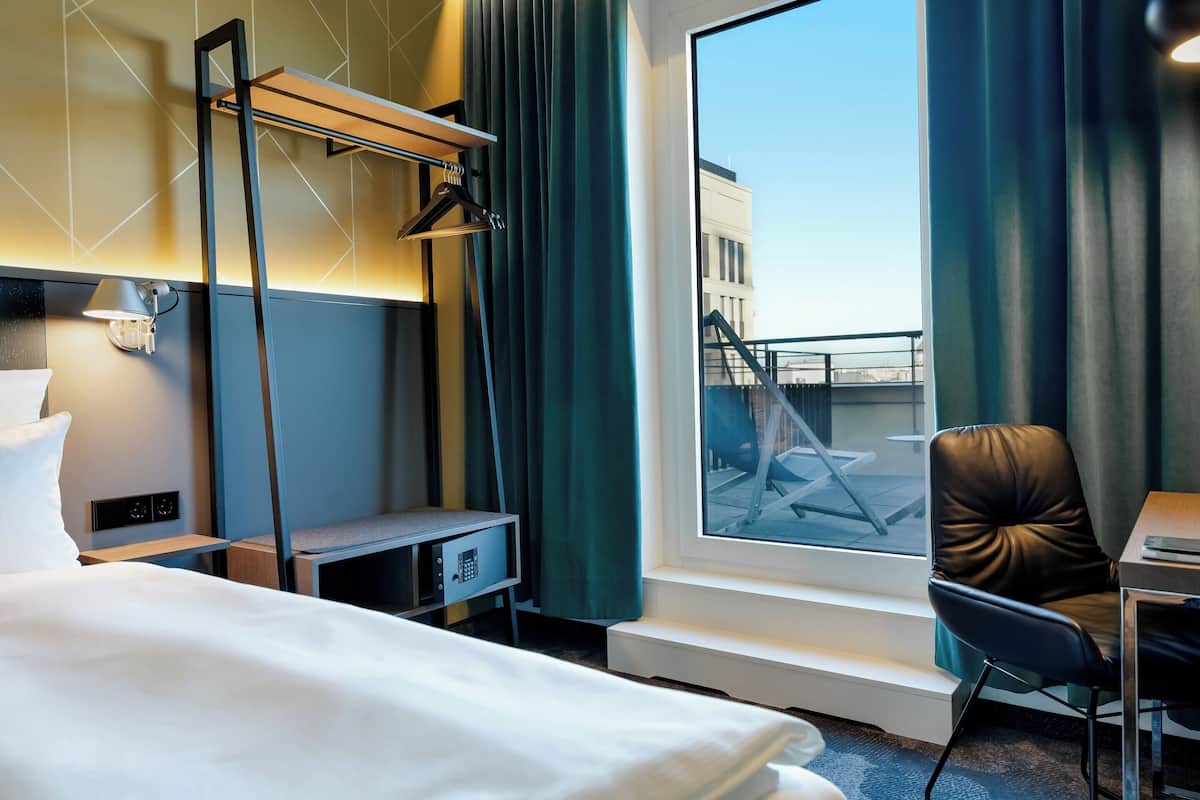 A hotel room with a bed, a desk with a chair, a desk lamp, a window with curtains, and a view of a building outside.