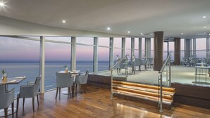 Un comedor moderno con grandes ventanales con vistas al mar, suelo de madera y mobiliario contemporáneo.