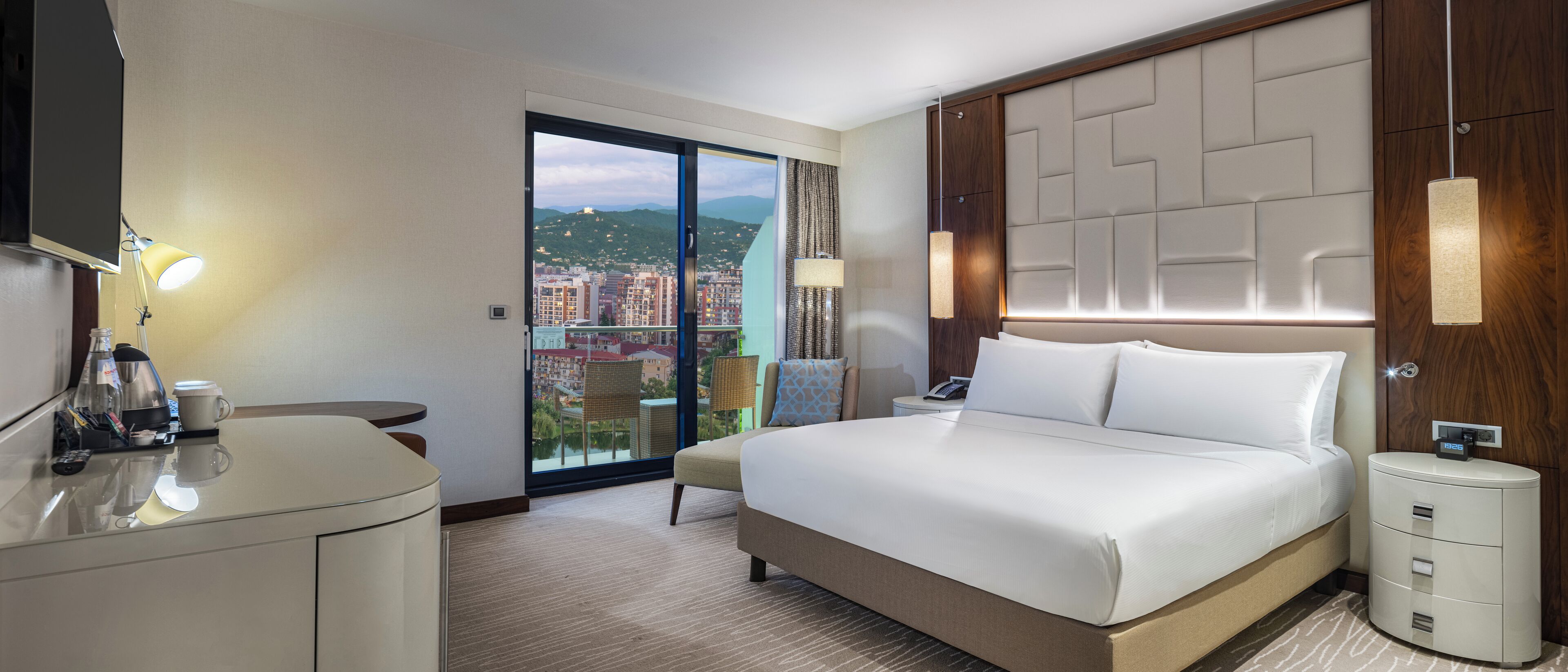 A hotel room with a large bed, a desk with a lamp, a chair, and a view of a cityscape through a sliding door.