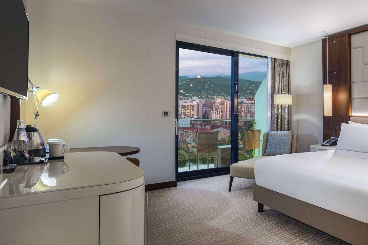A hotel room with a large bed, a desk with a lamp, a bedside table, and a view of a cityscape through a sliding door.