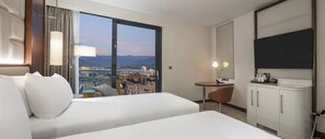 A hotel room with two beds, a desk, a chair, a TV, and a large window with a city view. - Hilton Batumi (Batumi)