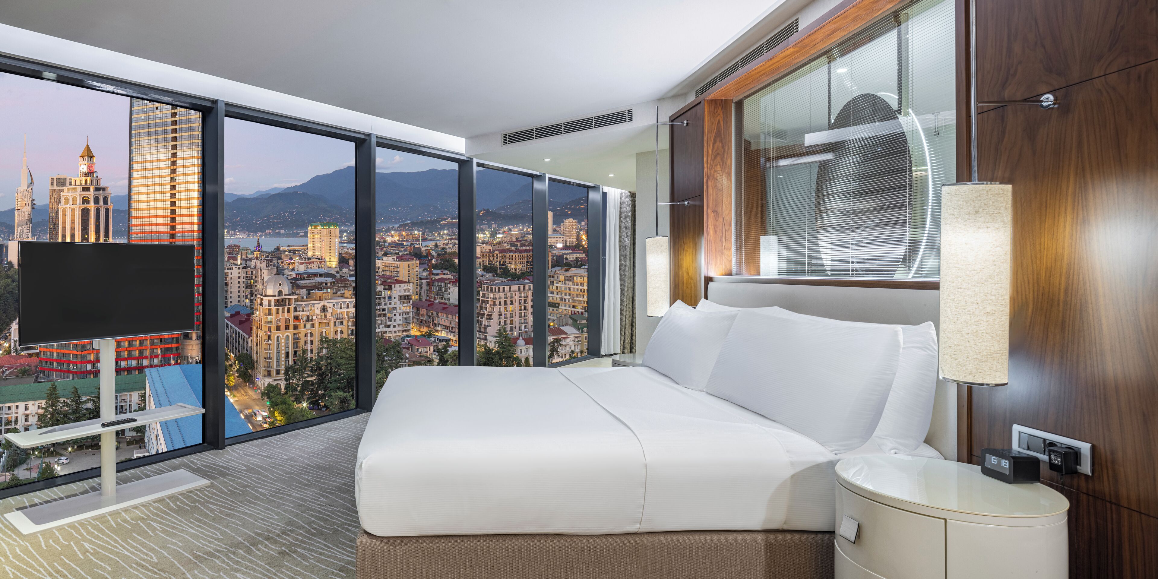 A modern hotel room with a large bed, a TV, a nightstand, and a view of a cityscape through floor-to-ceiling windows.