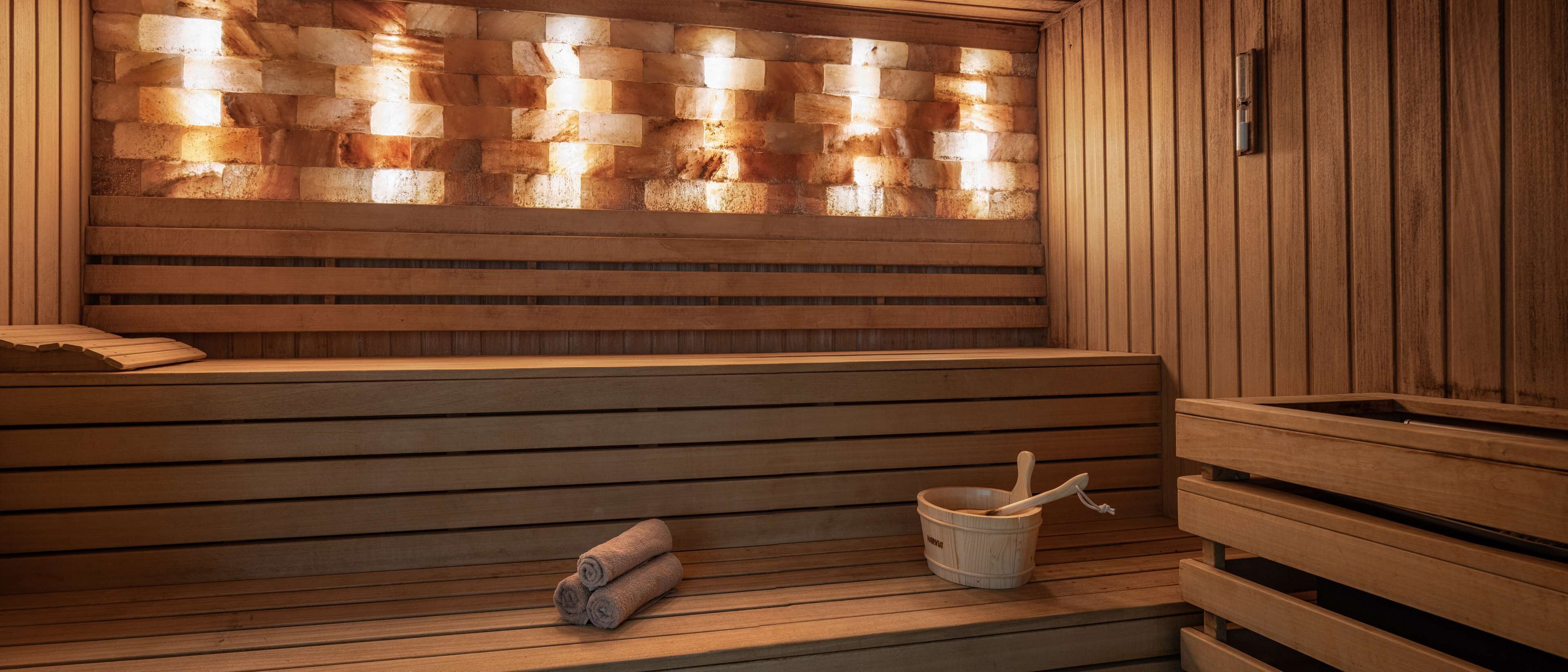 Un sauna en bois avec des bancs, un mur fait de blocs de pierre et un seau en bois avec une brosse.