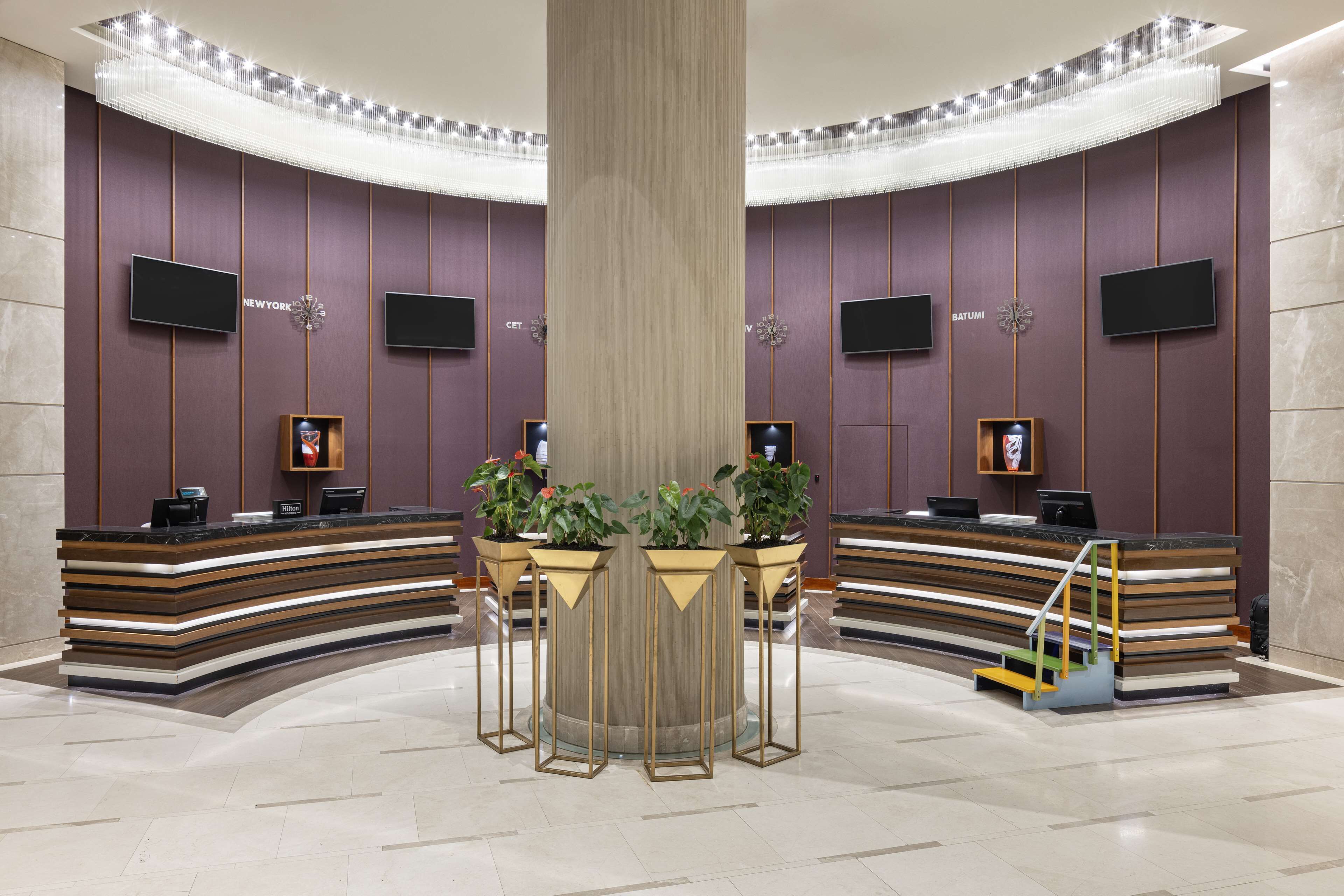 A reception area with a curved desk, two monitors, potted plants, and a central pillar.