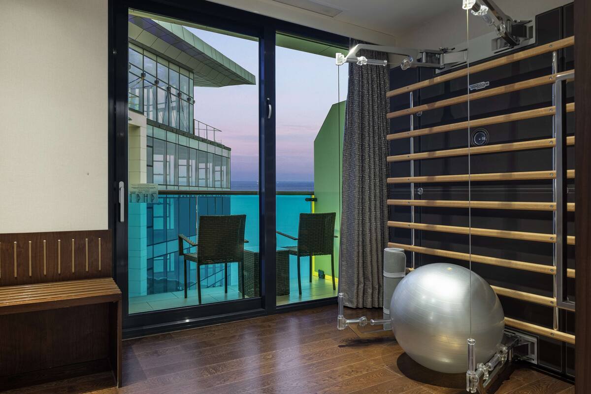 A room with a large window, a wooden bench, a pull-down exercise ball, and a wall-mounted rack.