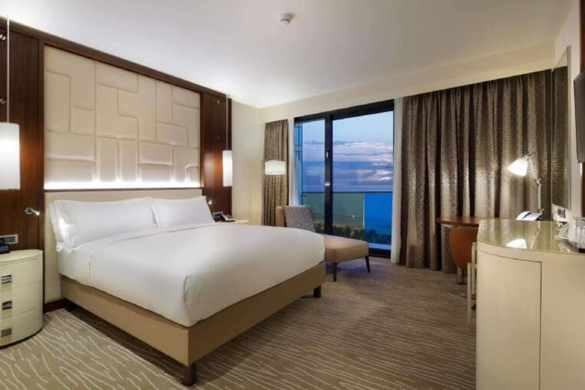 A hotel room with a large bed, a desk with a television, a chair, a nightstand, and a view of the outdoors.