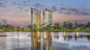 Un hotel moderno de muchos pisos, con su reflejo en el agua, y un parque con árboles y un lago al frente.