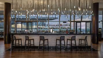 Una zona de bar con suelo de madera, cuatro taburetes, una barra con botellas y vistas al mar a través de amplios ventanales.