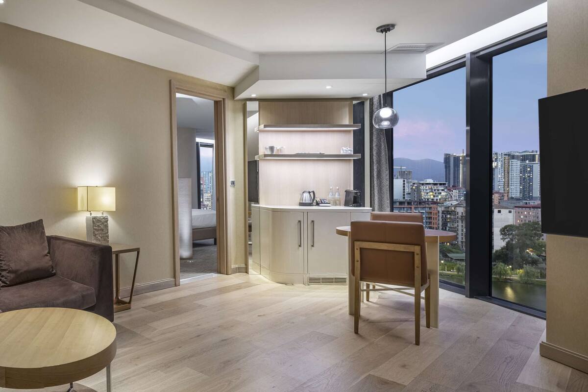 A modern living room with a sofa, a round coffee table, and a view of a cityscape through large windows.