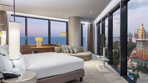 Una habitación de hotel moderna con una cama grande, un sofá, un escritorio y una televisión. La habitación tiene ventanas amplias con vistas a la ciudad.