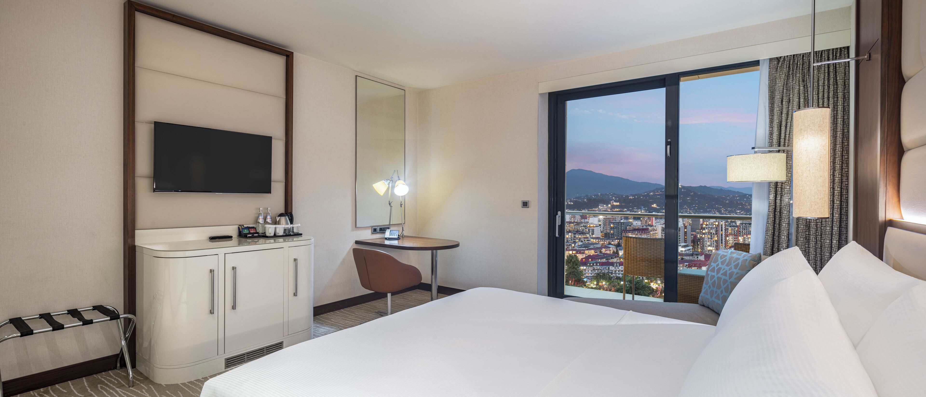 Habitación de hotel con cama, escritorio, silla, televisor, minibar y un amplio ventanal con vistas a la ciudad.