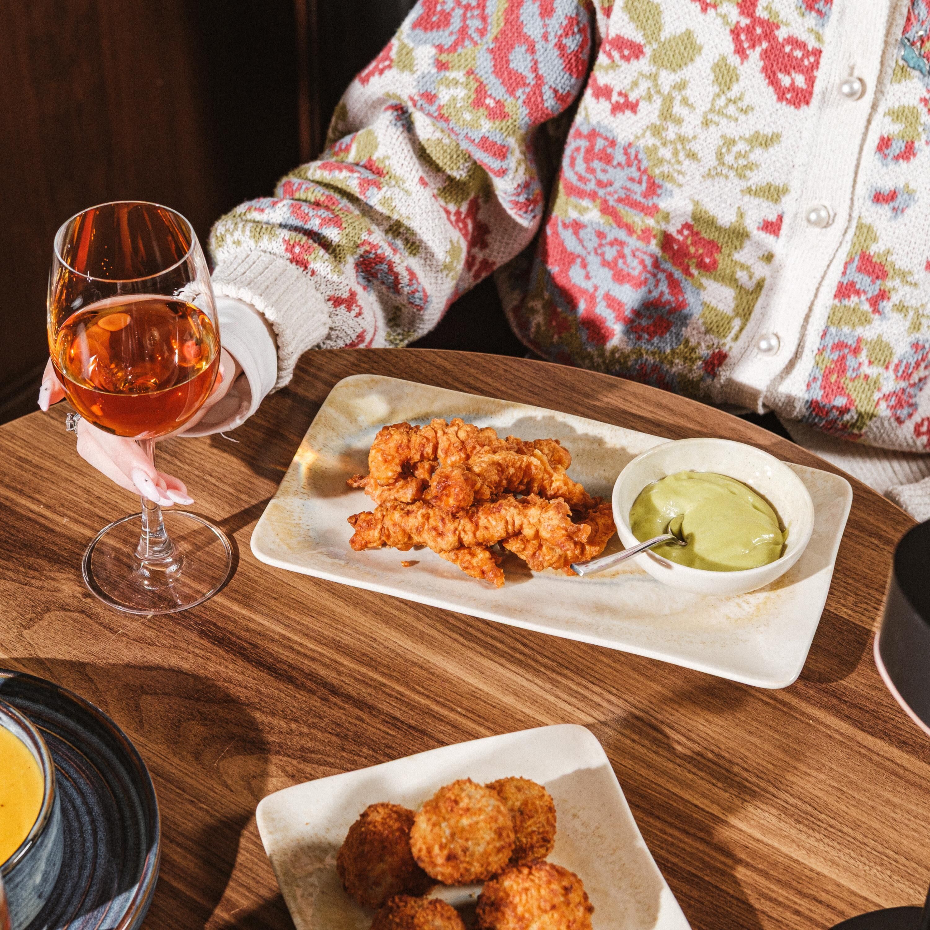 A dining table with a plate of fried chicken, a glass of amber-colored beverage, and a bowl of green sauce.