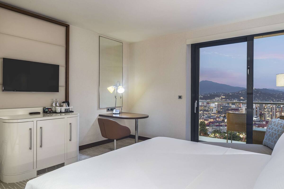 A hotel room with a bed, a desk, a chair, a TV, a mirror, a window with a city view, and a lamp.
